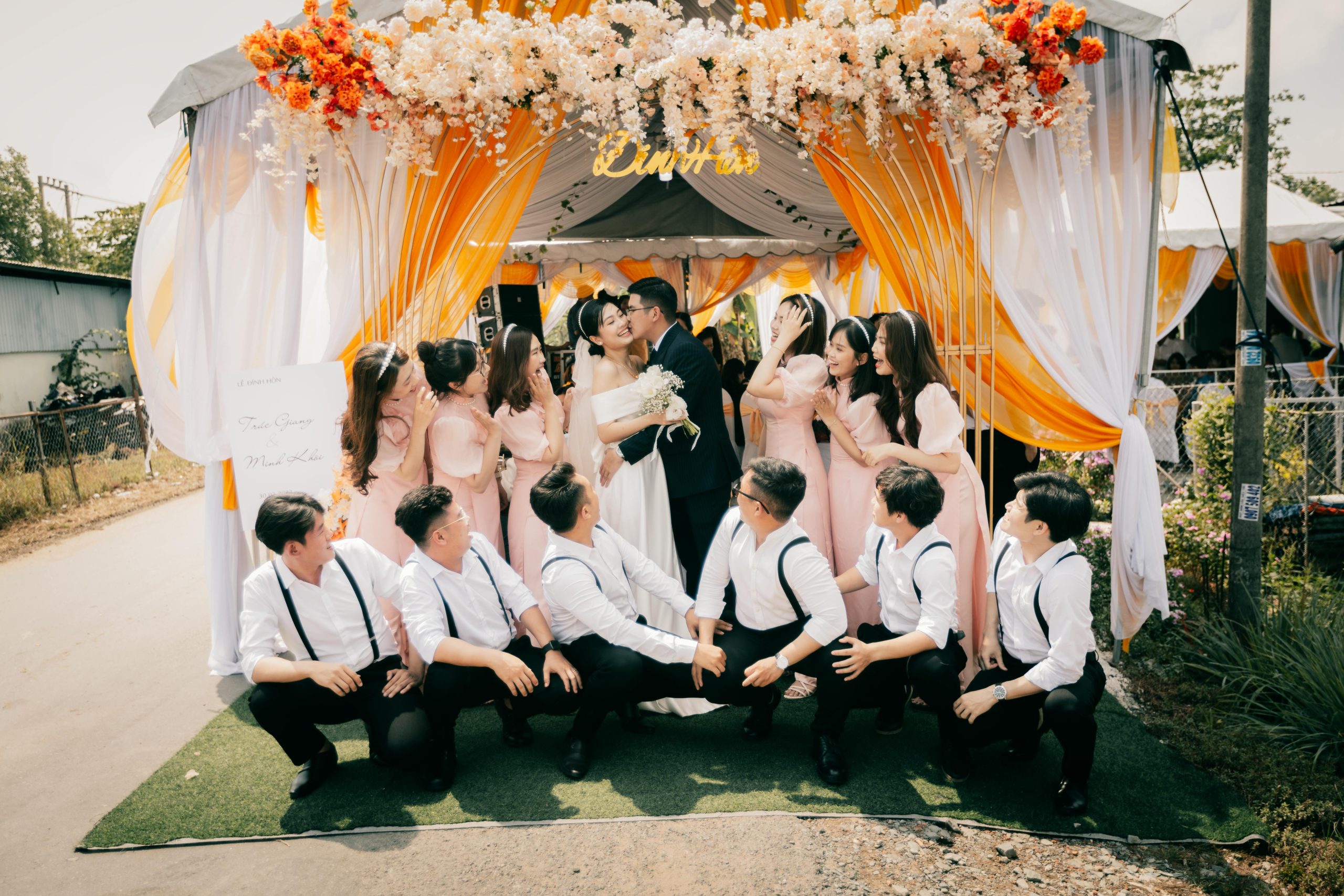 A joyful outdoor wedding ceremony with bride, groom, and bridal party under a floral tent display.